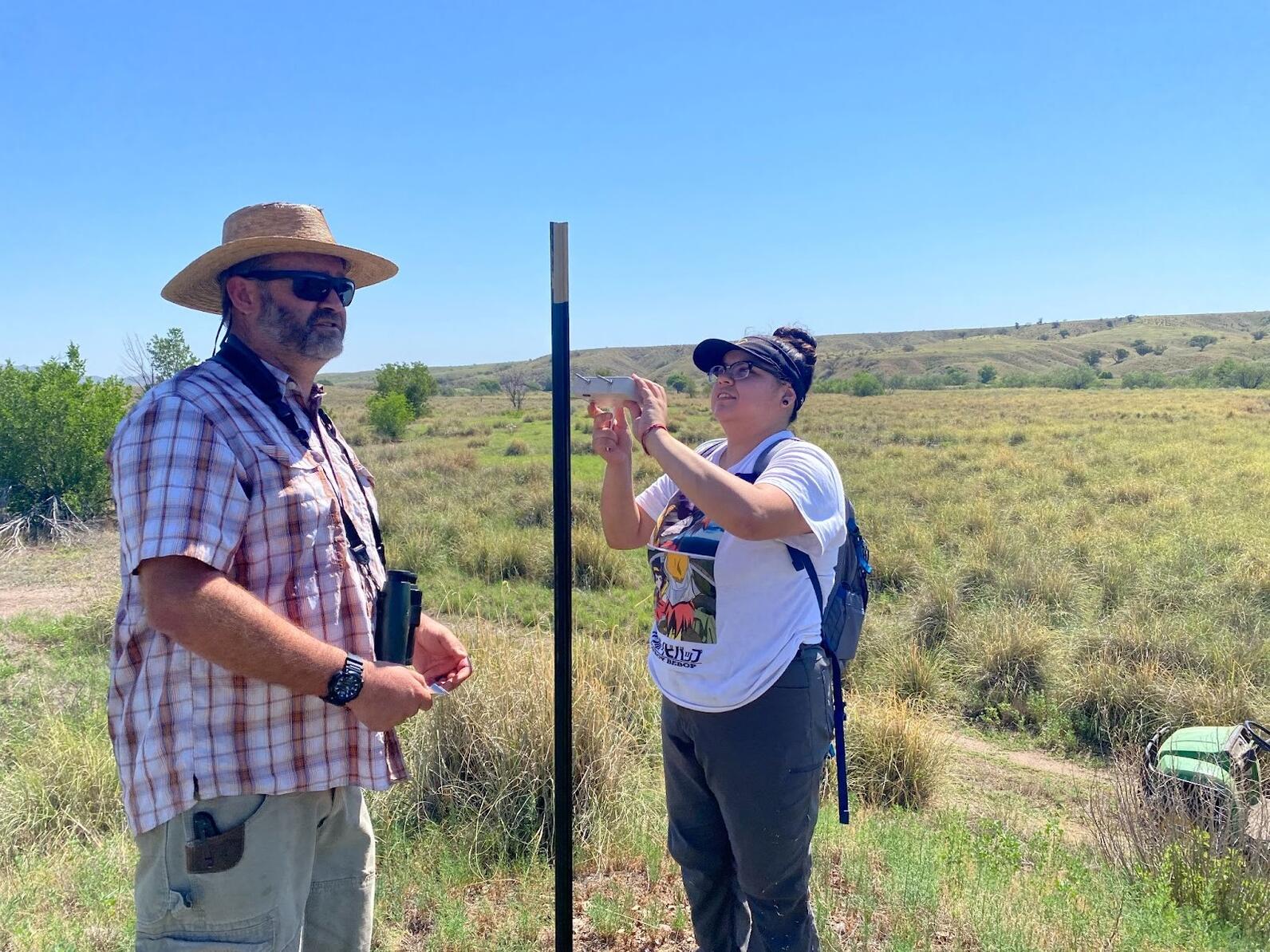 Heaven Perez Sanchez attaches a small, white box with a long antenna to a tall, green post as another field biologists stands by.