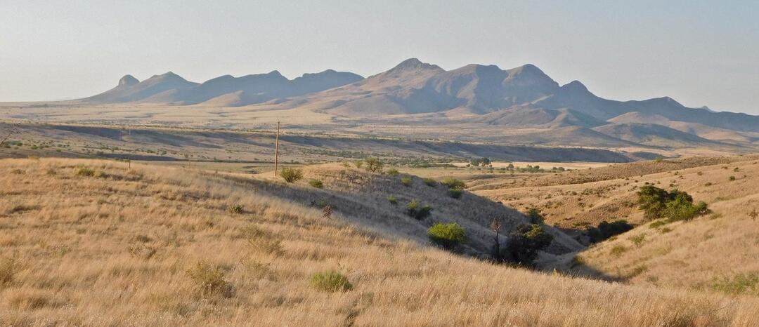 A rugged, isolated mountain range rises from a vast grassland against clear, blue skies.